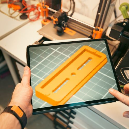 A person uses a tablet to view a 3D model prototype in a workshop equipped with 3D printers.